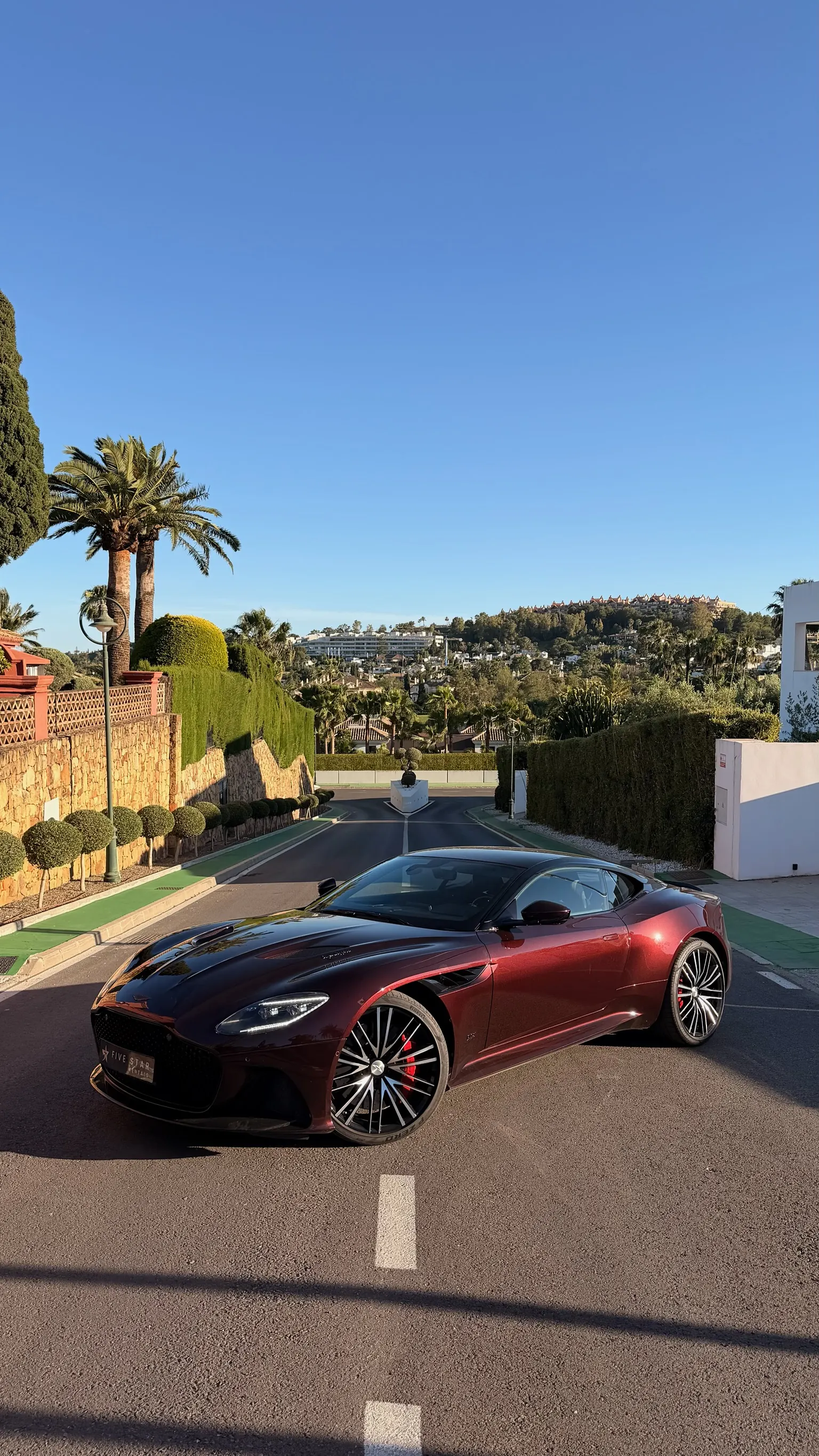 Aston Martin DBS Superleggera in Divine Red, front three-quarter view with Five Star Rentals plate, 21-inch Y-Spoke wheels and Marbella panorama