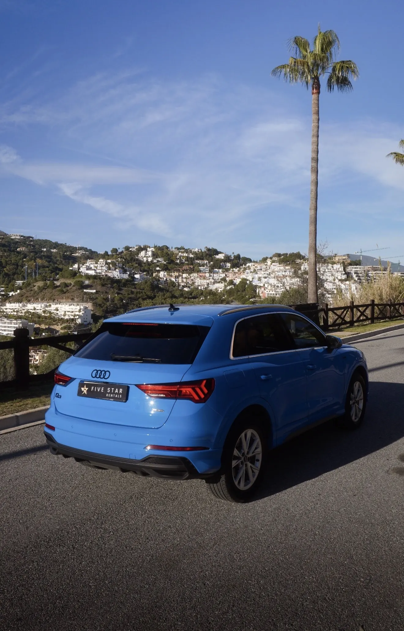 Rear three-quarter view of the Turbo Blue Audi Q3 with Five Star Rentals plate, dynamic LED tail lights and white hillside village of Marbella in background