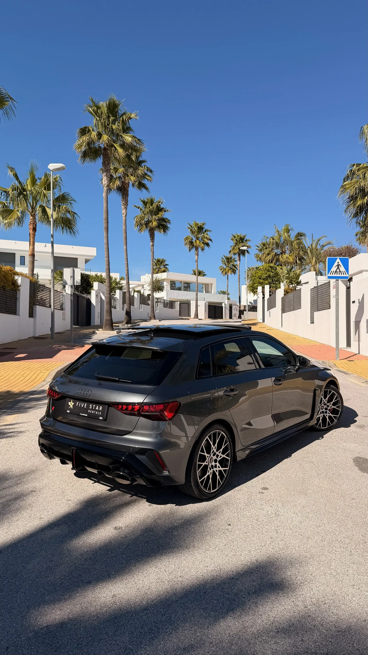 Rear three-quarter view of the grey Audi RS3 Sportback with row of Marbella palm trees and glass panoramic roof