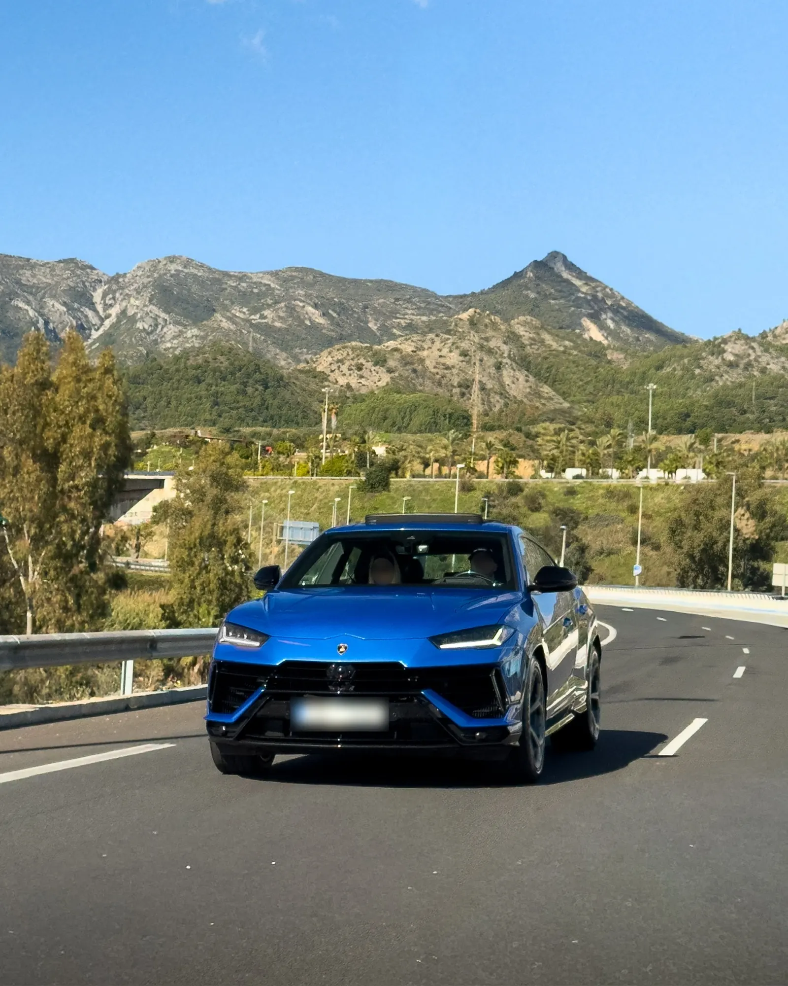 Lamborghini Urus S in Blu Eleos driving front view with hexagonal grille, Y-shaped LED headlights and Marbella mountains in background