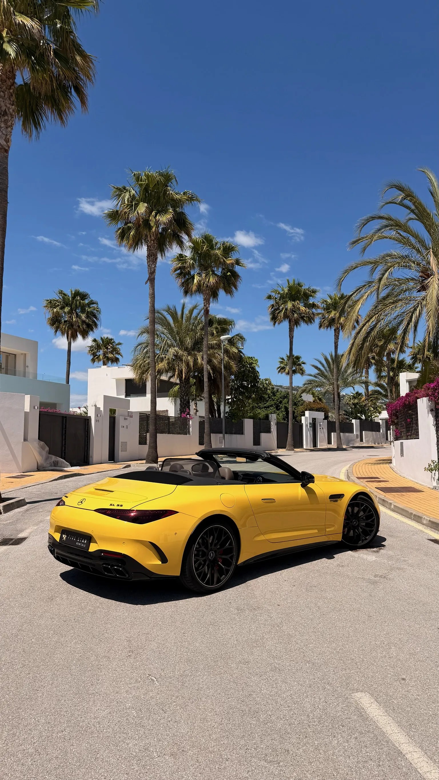 Rear three-quarter view of the Sun Yellow Mercedes-AMG SL 55 with top down, Five Star Rentals plate, Marbella palm trees and bougainvillea in background