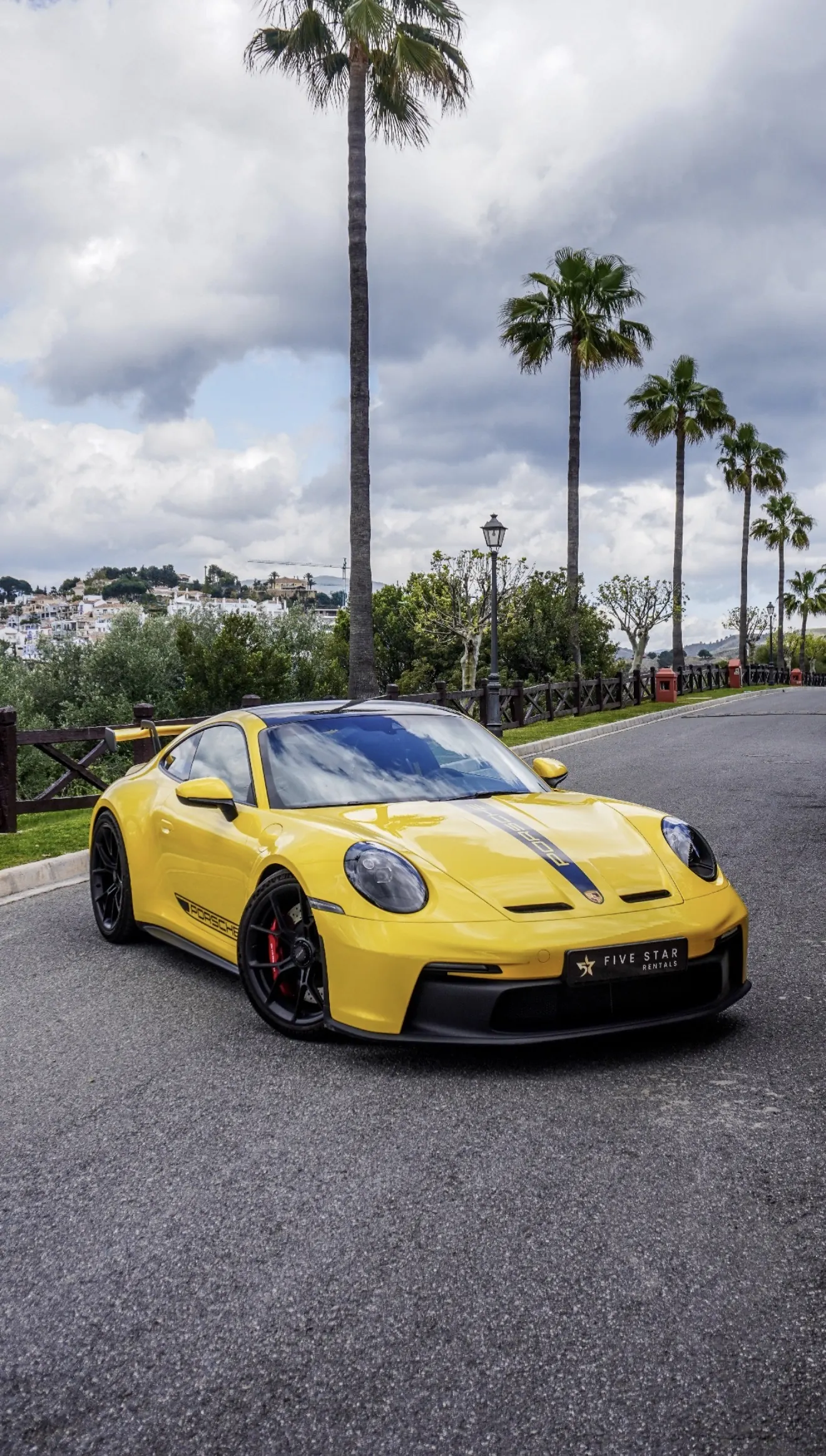 Porsche 911 GT3 type 992 in Racing Yellow, front three-quarter view with Five Star Rentals plate, Marbella palm trees and white village in background