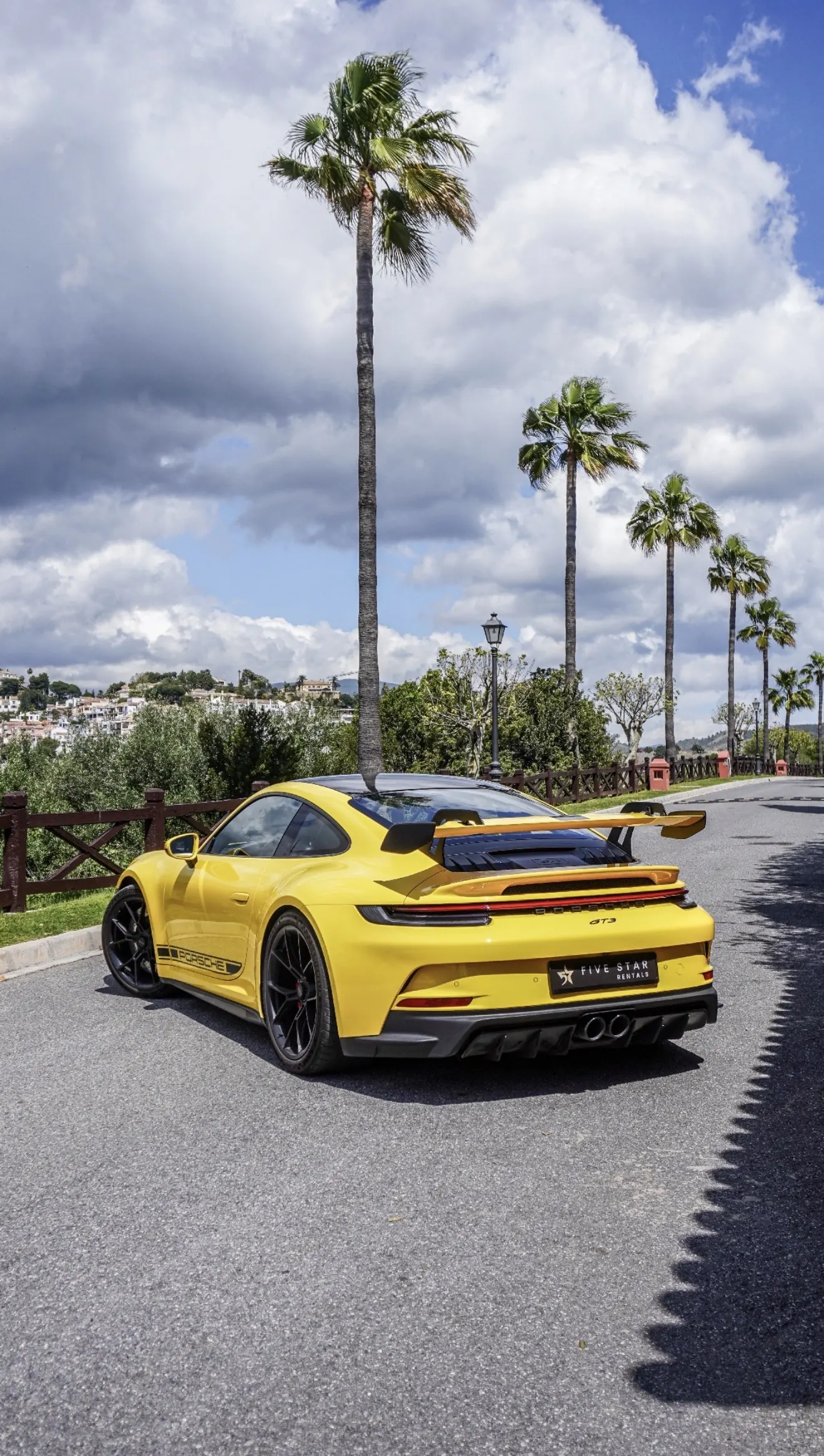 Rear three-quarter view of the Racing Yellow Porsche 911 GT3 992 with swan-neck rear wing, GT3 badge and Five Star Rentals plate under Marbella palm trees