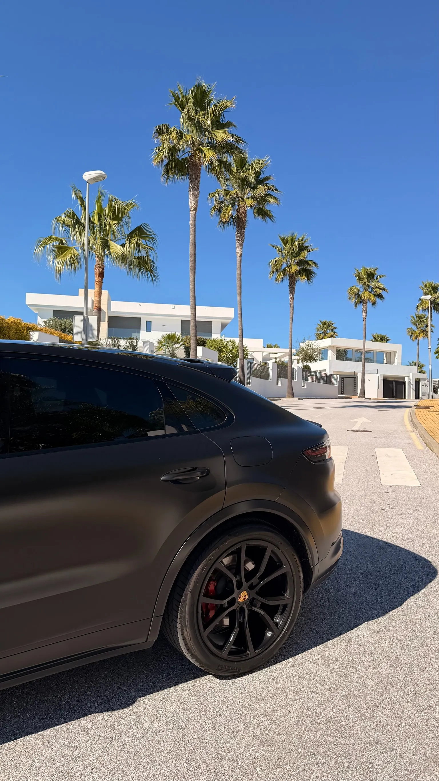 Rear side profile of the matte black Porsche Cayenne Coupé with close-up black wheel and Marbella palm trees